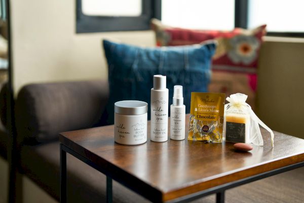 The image shows spa products on a table: body butter, body oil, mist, chocolate, soap, with colorful cushions in the background.