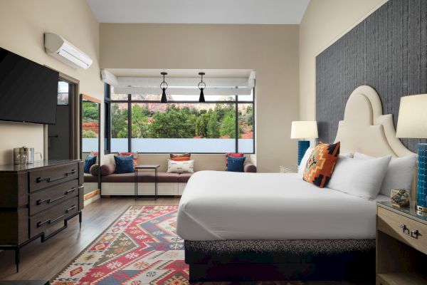 A modern hotel room with a large bed, colorful rug, window seating, and a view of nature outside.