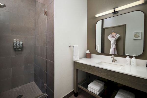 A modern bathroom featuring a shower, a vanity with a sink, mirror, towels, and toiletries. A robe hangs in the reflection.