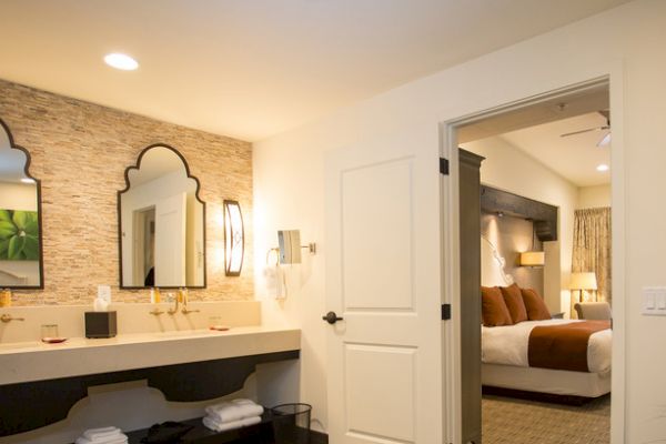 The image shows a well-lit hotel room with a bathroom featuring a double sink and ornate mirrors, leading into a bedroom with a bed and nightstand.