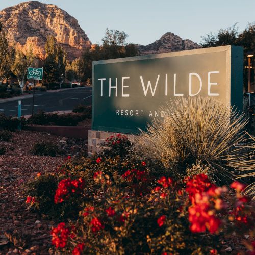 The image shows a sign for "The Wilde Resort and Spa" with vibrant red flowers in the foreground and scenic mountains in the background, under a clear sky.