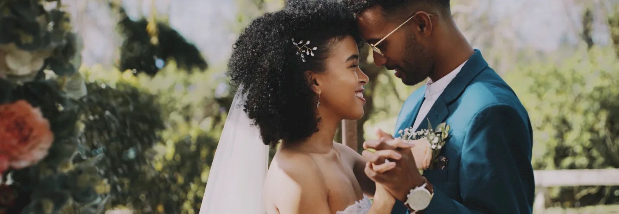 A bride and groom sharing a dance outdoors, surrounded by greenery and flowers, both smiling and embracing each other tenderly.
