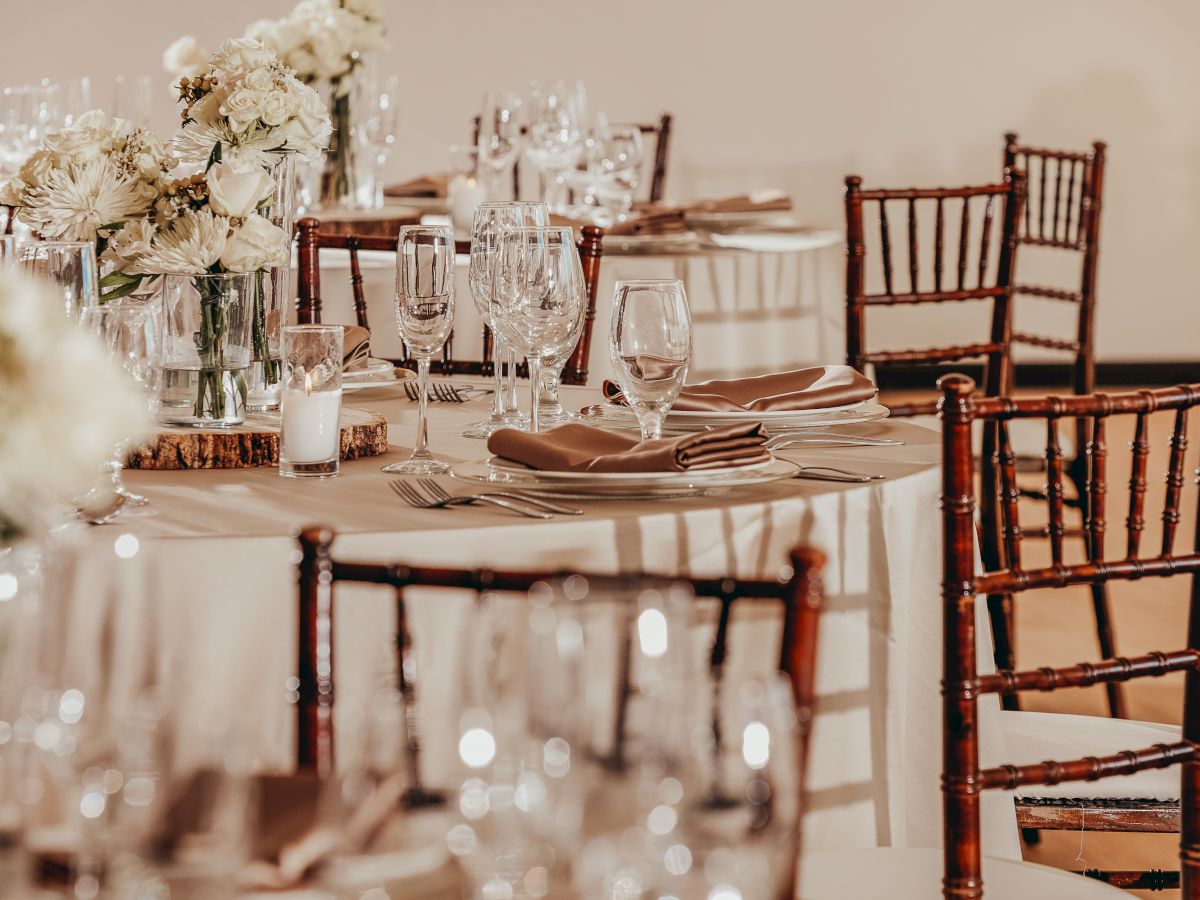 The image shows elegantly set round tables with white tablecloths, floral centerpieces, glassware, plates, napkins, and brown wooden chairs.