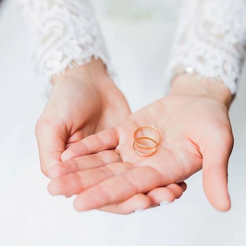 The image shows a person in white lace sleeves holding two gold rings in their cupped hands, likely a bride on her wedding day.