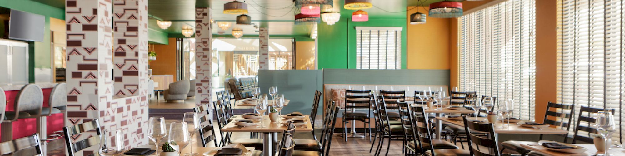 A brightly lit, colorful restaurant with eclectic decor, featuring multiple tables and chairs, vibrant ceiling lights, and large windows on the right.