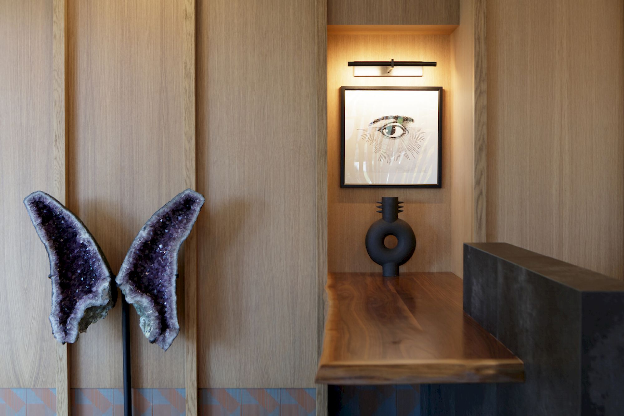 The image shows a wooden wall with butterfly-shaped crystals, a lighted framed eye artwork, a circular sculpture, and a wooden table/desk.