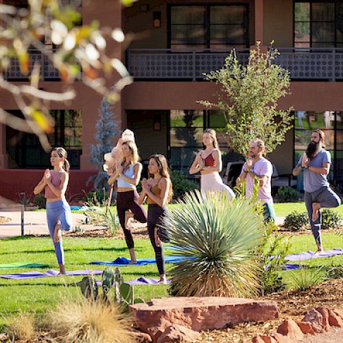 A group of people are practicing outdoor yoga on mats in a garden area, with buildings and greenery in the background.