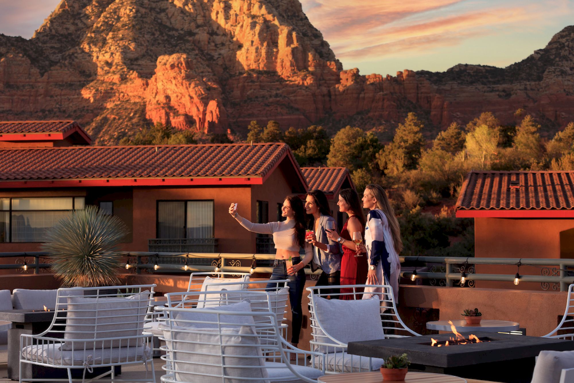 A group of people are taking a selfie on a rooftop with a scenic mountain backdrop, surrounded by outdoor seating and warm evening light.