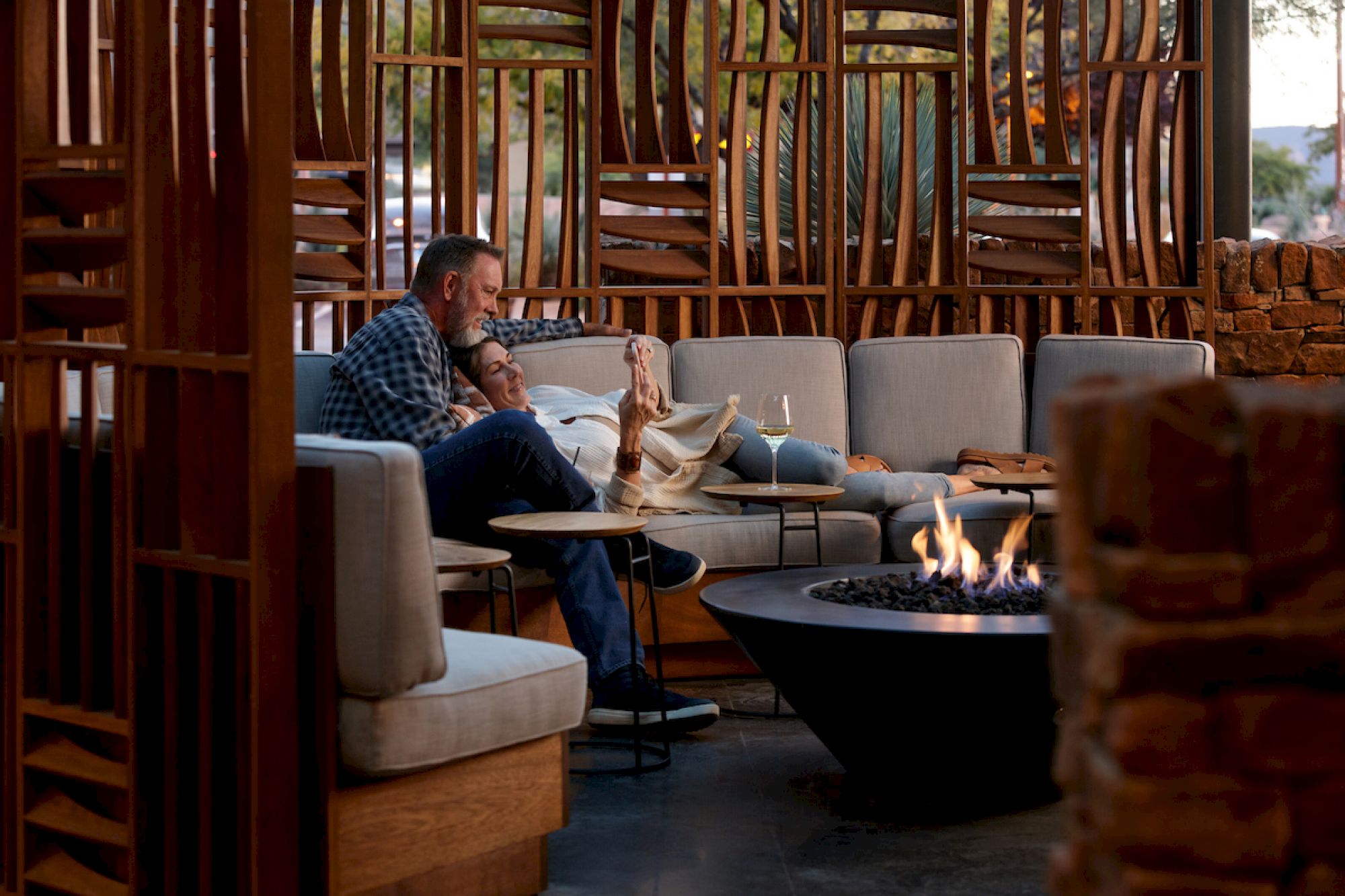 A couple relaxes on a modern outdoor sofa by a fire pit, surrounded by wood paneling and decor, creating a cozy atmosphere.