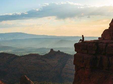 A person sits on a rocky cliff's edge, overlooking a vast landscape with mountains and a cloudy sky during sunset.
