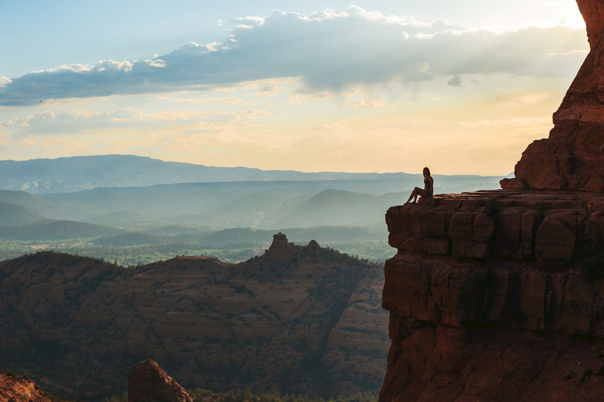 A person sits on the edge of a rocky cliff, overlooking a vast mountainous landscape under a partly cloudy sky during sunset.
