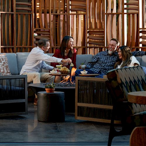 Four people are seated around a fire pit in a cozy setting with wooden decor, enjoying food and drinks, and engaging in conversation.