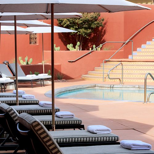 A serene poolside scene with sun loungers, umbrellas, and a staircase surrounded by a vibrant red wall and cacti, ready for relaxation.