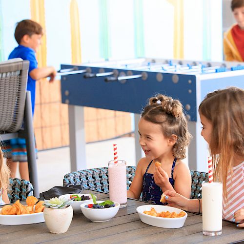 Children enjoying snacks and drinks at a table, with some playing foosball in the background. The atmosphere is bright and cheerful.