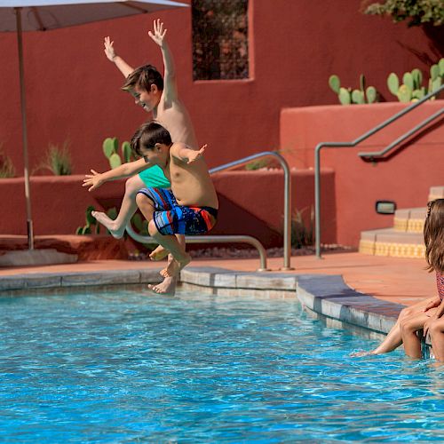 Two boys are jumping into a pool while two girls sit at the edge, dipping their feet in the water. The background shows steps and a red wall.