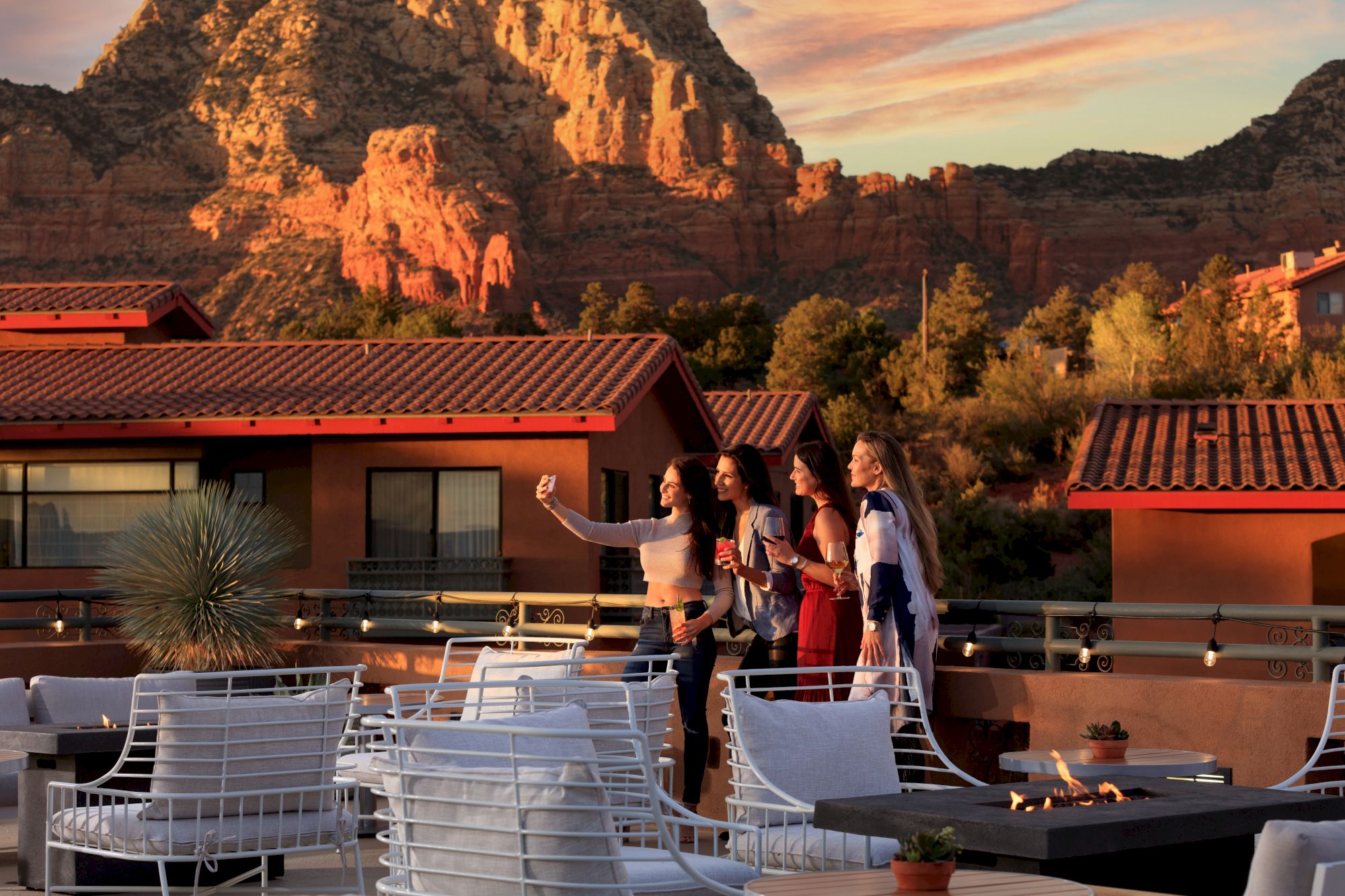 A group of people is taking a selfie on a rooftop patio with a scenic background, including mountains and buildings, during sunset.