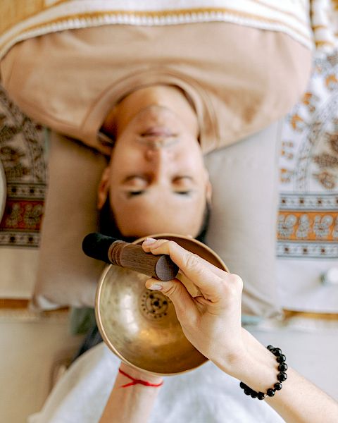 A person is lying down with a singing bowl being used above them. Other bowls and a patterned cloth are visible around the person.