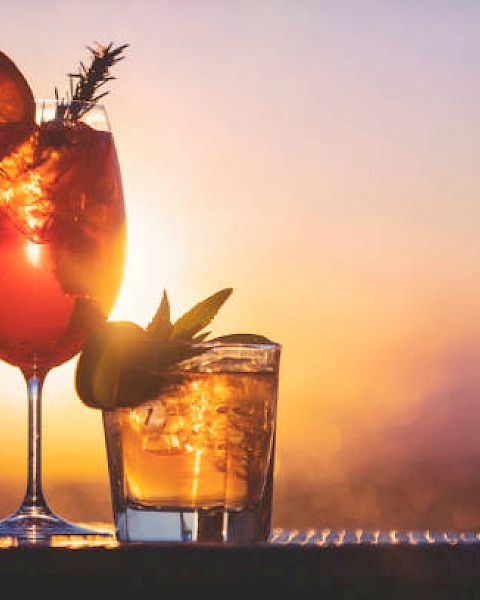 Two cocktails with ice and citrus garnishes, set against a vibrant sunset background.