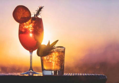 Two cocktails with ice and citrus garnishes, set against a vibrant sunset background.