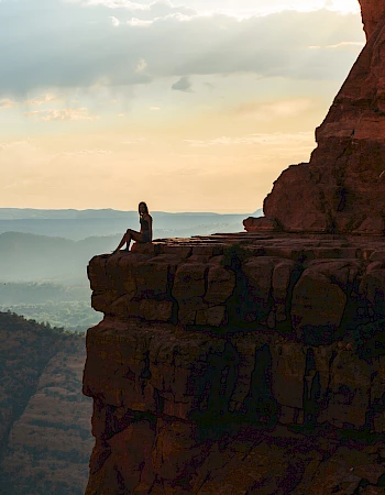 A person sits on the edge of a cliff at sunset, overlooking a vast, misty landscape with layered hills in the distance.