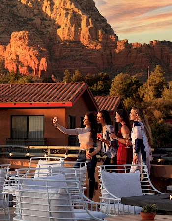 A group of people taking a selfie on a rooftop with a mountain backdrop, surrounded by outdoor seating and warm lighting at sunset.