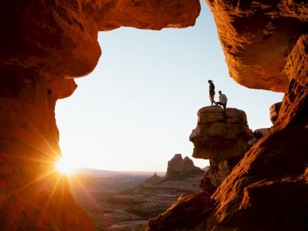 Silhouettes of two people on a rock formation during sunset, framed by surrounding cliffs, creating a dramatic landscape scene.