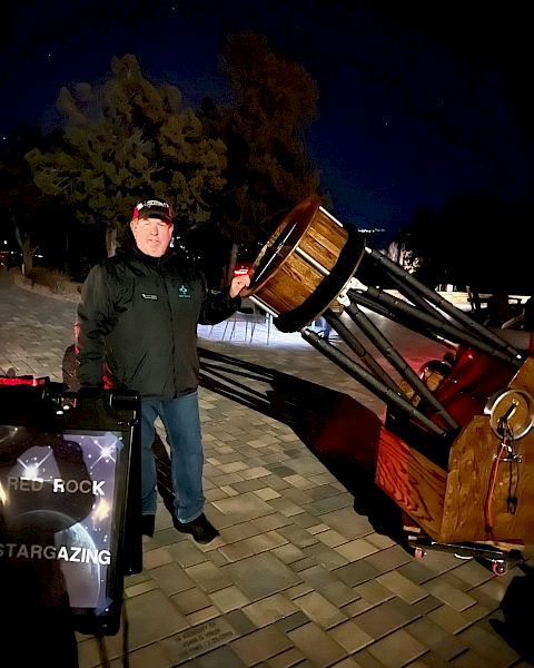 A person stands outside at night beside a large telescope, with a sign that reads "RED ROCK STARGAZING." Trees and a dark sky are in the background.
