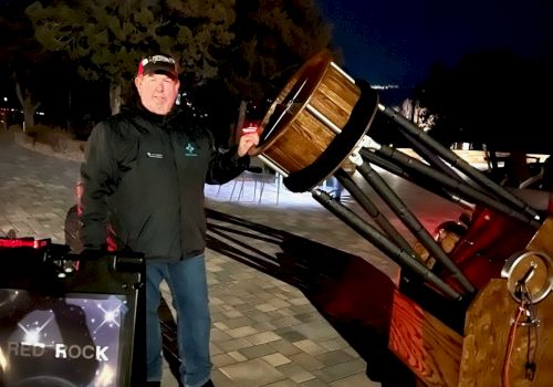 A person stands outside at night beside a large telescope, with a sign that reads "RED ROCK STARGAZING." Trees and a dark sky are in the background.