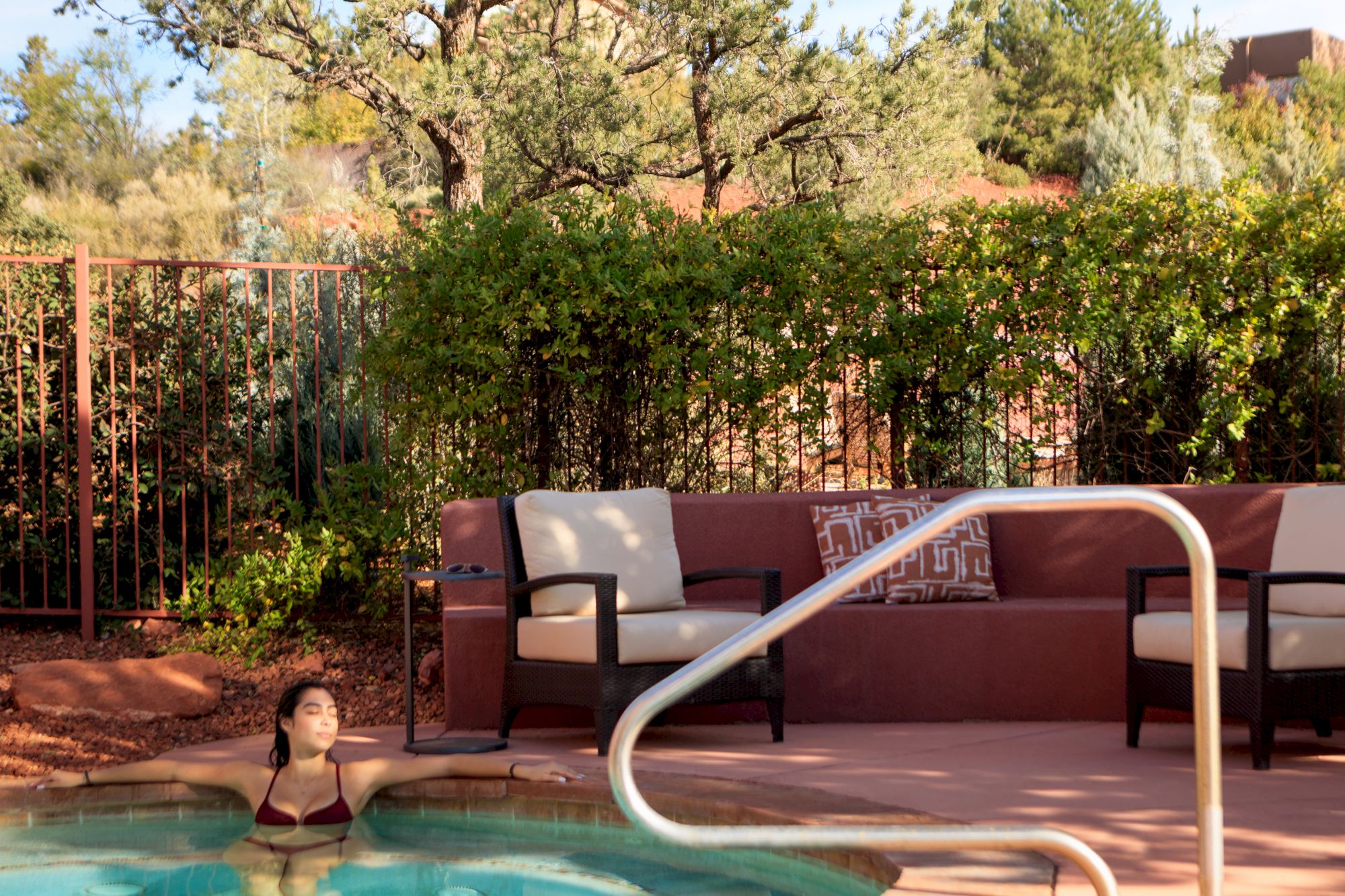 A person in a red bikini is relaxing in a hot tub; there are chairs and greenery in the background.