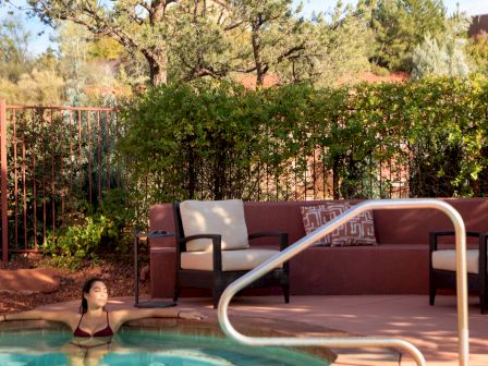 A person relaxes by a pool surrounded by chairs and greenery, with a scenic outdoors background under a clear sky.