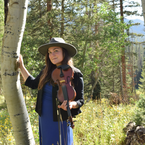 A person in a hat stands in a forest with a violin, leaning against a tree amidst greenery and sunlight filtering through the trees.