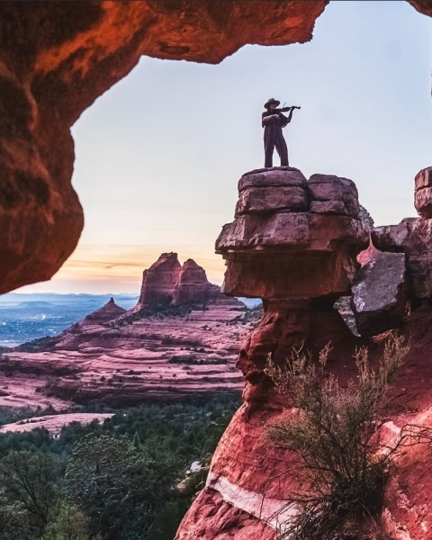 A person stands on a rock formation, playing a flute, surrounded by red sandstone cliffs and a scenic desert landscape at sunset.