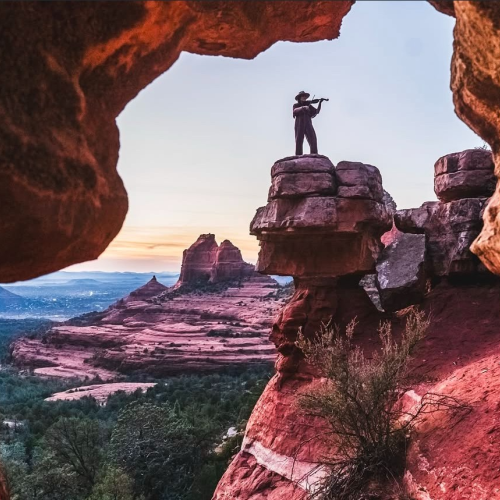 A person stands on a rock formation, playing a flute, surrounded by red sandstone cliffs and a scenic desert landscape at sunset.