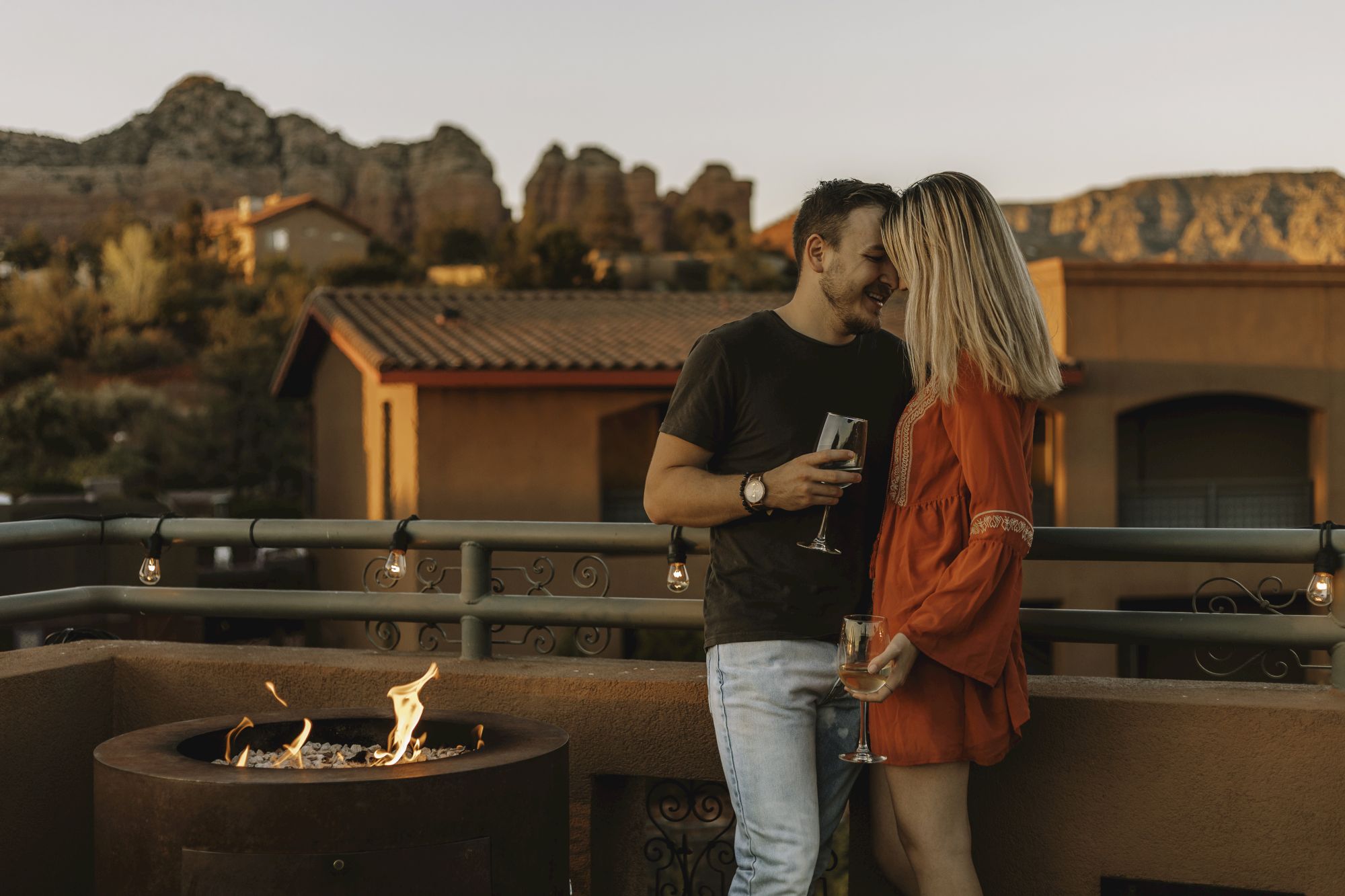 A couple stands by a fire pit on a patio, holding glasses, with scenic mountains and a house in the background.
