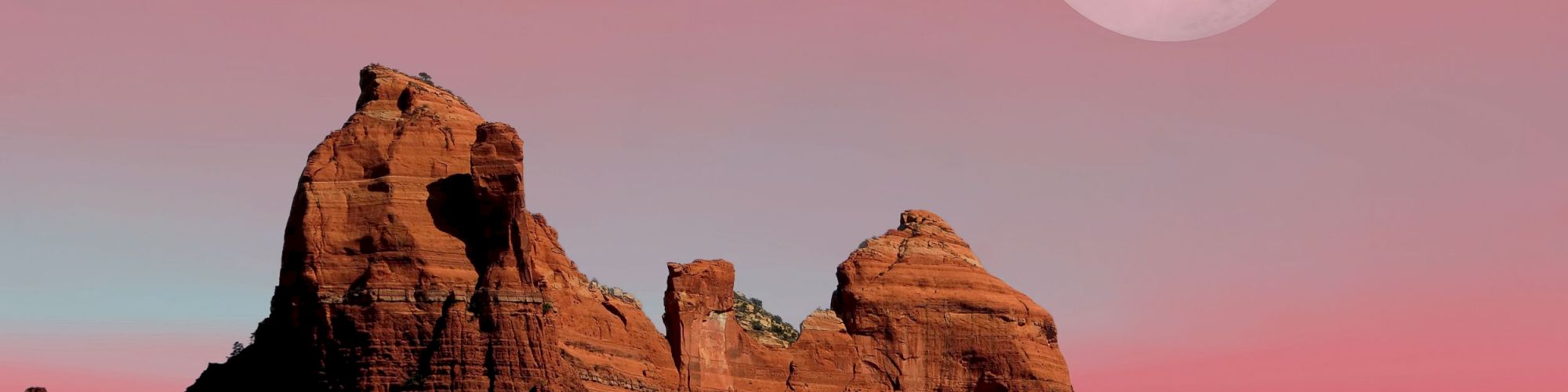 A reddish rock formation under a pink sky with a large, bright moon above; the landscape features desert vegetation.