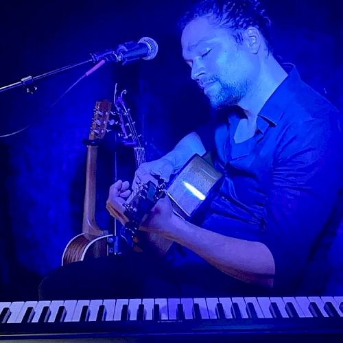 A person is playing guitar under blue lighting, with a microphone and keyboard nearby, creating a musical atmosphere.