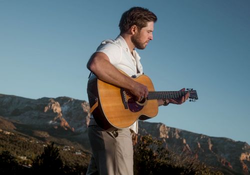 A man is playing an acoustic guitar outdoors, with a mountainous landscape in the background and clear skies above.